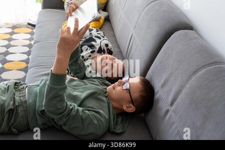 Two brothers lying on grey sofa watching video on smartphone together with happy smiles at home. Concept of siblings bonding, mobile entertainment, di Stock Photo