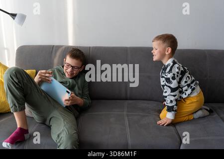 Two brothers laughing on grey sofa with older boy holding tablet while younger sibling watches happily at home. Concept of siblings playful interactio Stock Photo