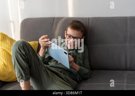 Happy boy with glasses playing game on tablet while relaxing on grey sofa with yellow pillow at home. Concept of children screen time entertainment, d Stock Photo