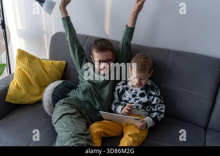 Excited boy with raised arms celebrating while younger brother holds tablet on grey sofa at home. Concept of gaming victory, siblings digital fun, chi Stock Photo