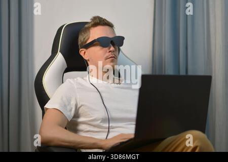 Relaxed man sitting in a chair and using wearable AR XR smart glasses while working on a laptop. Modern technology, virtual reality and digital lifest Stock Photo