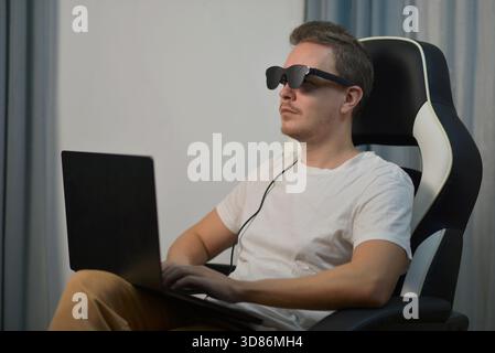 Relaxed man sitting in a chair and using wearable AR XR smart glasses while working on a laptop. Modern technology, virtual reality and digital lifest Stock Photo