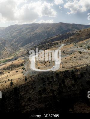 Aerial view of a winding road cutting through the rugged, sun-drenched mountains under a sky brushed with clouds, Vardenyats Pass, Armenia. Stock Photo