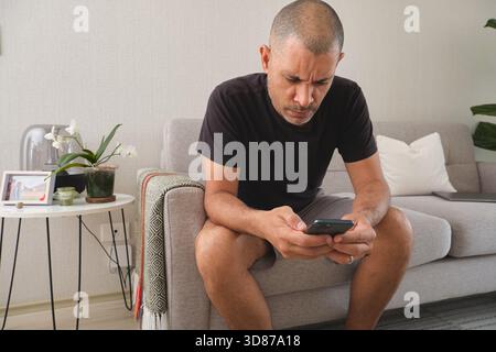 Man with a furrowed brow focusing on his mobile phone while sitting on a sofa Stock Photo