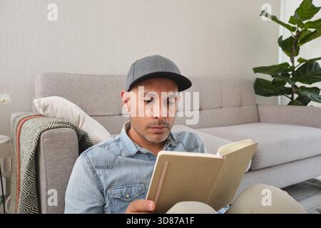 Man focused, sitting on floor in living room, reading a book Stock Photo