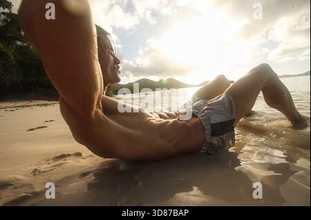 Close-up view of a man doing an abs workout on the beach during a golden sunset. Masculine sporty model is doing sit-ups, The scene shows exercise. Stock Photo