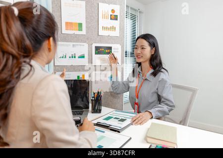 Two Asian businesswomen discussing company strategy together in a modern office, analyzing financial documents and charts. They are collaborating and Stock Photo