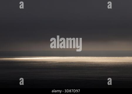 Sunlight breaking through clouds over dark ocean horizon Stock Photo