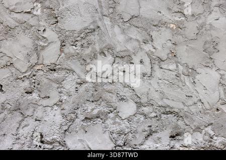 Rough textured concrete wall displayed in outdoor setting. Daylight illuminates surface, highlighting neutral tones and rugged features of the wall. Stock Photo