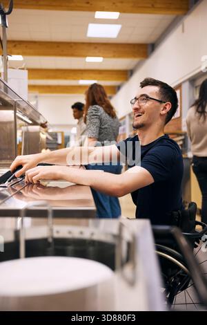Happy young man taking food from container while sitting on wheelchair in school cafeteria Stock Photo