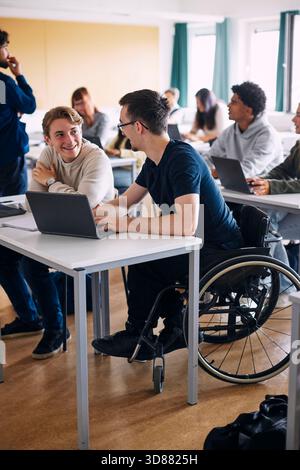 Young man sitting on wheelchair talking with male classmate while studying on laptop in high school classroom Stock Photo