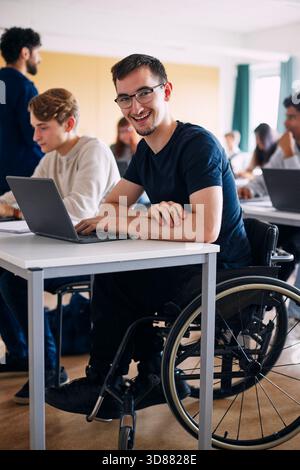 Portrait of physically disabled happy young man sitting with laptop at desk in high school classroom Stock Photo