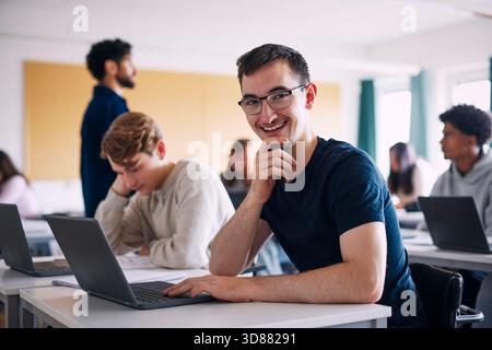 Portrait of physically disabled happy young man sitting with hand on chin at desk in high school classroom Stock Photo