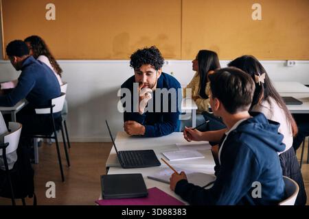Male teacher assisting high school students while leaning on desk in classroom Stock Photo