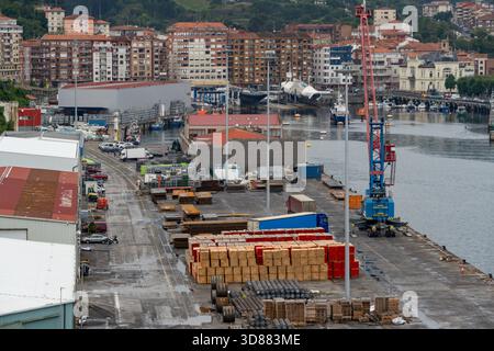 Industrial Cargo Dock at Bermeo Port, Stacks of Timber and Metal, Crane, Trucks, Warehouses, and Residential Buildings Background, Basque Country Stock Photo