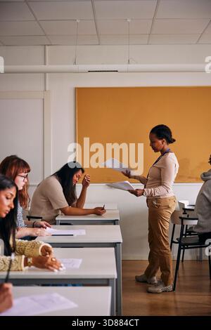Female teacher giving test paper to teenage student sitting at desk in high school classroom Stock Photo