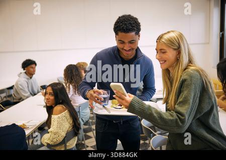 Smiling blond girl showing smart phone to male friend standing with food plate during lunch break in high school cafeter Stock Photo