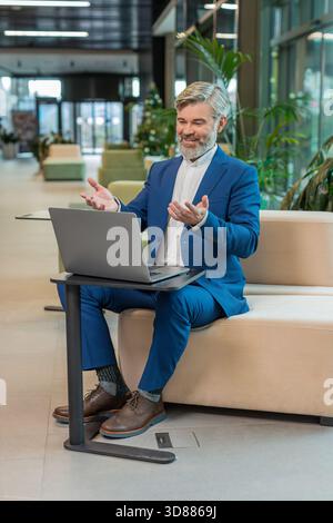 Middle-aged businessman working on laptop computer in modern office lobby talking on online communication video call with employee, boss. Manager man support services looking at webcam of computer. Stock Photo