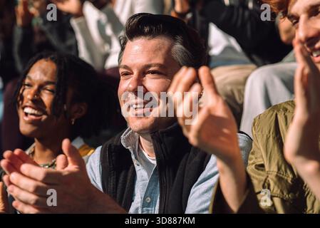 Smiling mature man clapping while sitting at sports stadium on sunny day Stock Photo