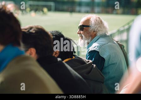 Side view of smiling senior man wearing sunglasses watching game while sitting with audience at sports stadium stand Stock Photo