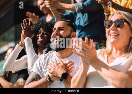 Happy bearded man celebrating with hands on chest surrounded by fans in stadium stand on sunny day Stock Photo