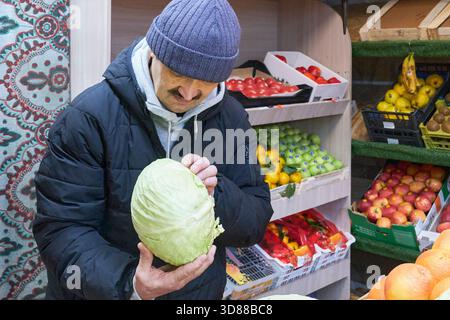 Senior man in warm jacket and knit cap holds and examines head of light green cabbage while shopping for vegetables at market. Shelves stocked with fr Stock Photo