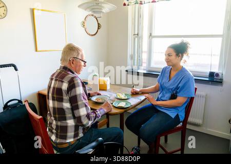 Female caregiver sitting with senior man eating breakfast at home Stock Photo
