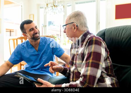 Elderly man holding digital tablet talking with male nurse while sitting on armchair at home Stock Photo