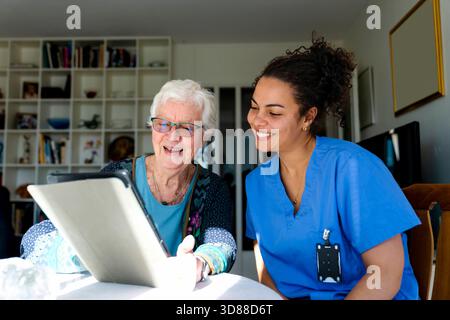 Smiling female healthcare worker sitting near senior woman using digital tablet while sitting at home Stock Photo