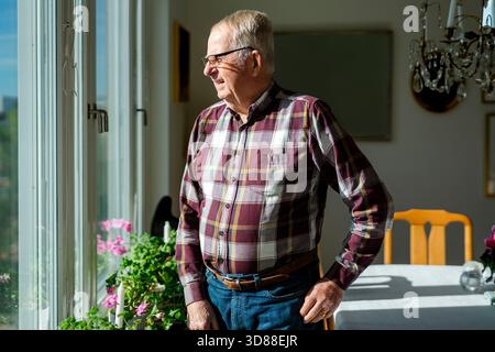 Senior man looking out window while standing in living room at home Stock Photo