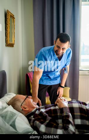 Happy senior man lying on bed while talking with male caregiver in bedroom at home Stock Photo