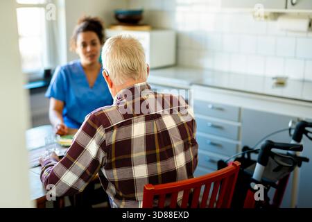 Rear view of elderly man sitting on chair while talking with female caregiver at home Stock Photo