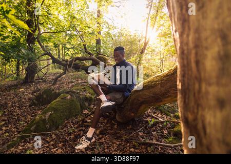 Man reading book while sitting on tree trunk in forest Stock Photo