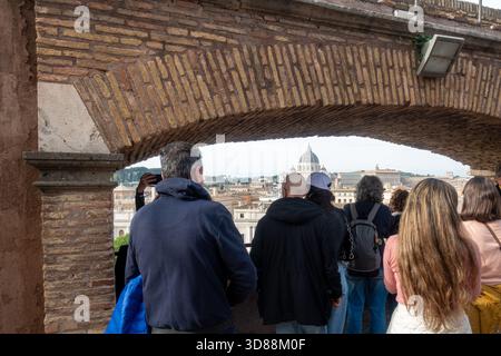 A group of tourists are looking at the view of Rome, Italy, through a brick archway. In the distance, St. Peter's Basilica can be seen. Stock Photo