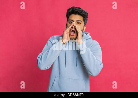 Excited Indian man shouting, holding hands near mouth, calling out loudly, waving and expressing urgency or enthusiasm. Energetic Arabian guy communicating with emotion, isolated on red background Stock Photo