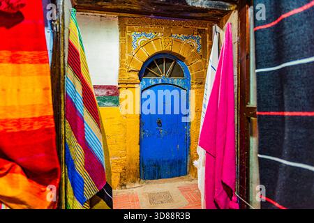 Ornate blue door with Islamic arch framed by yellow walls and colorful striped textiles in Essaouira medina, Morocco Stock Photo