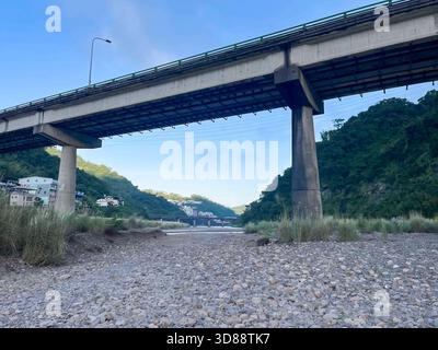 Looking up at the massive concrete structure of an elevated viaduct from the rocky riverbed. Green hills and village buildings visible in the distance Stock Photo