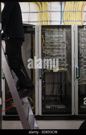 Detailed view of a modern server room with network cabinets full of organized fiber-optic and Ethernet cables. A technician standing on a ladder works Stock Photo