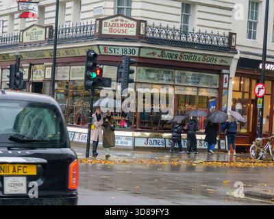 Exterior of James Smith and Sons, Grade II listed Victorian shop selling Umbrellas, parasols and walking sticks established in 1830, during rain storm with pedestrians passing holding umbrellas, Central London UK Stock Photo