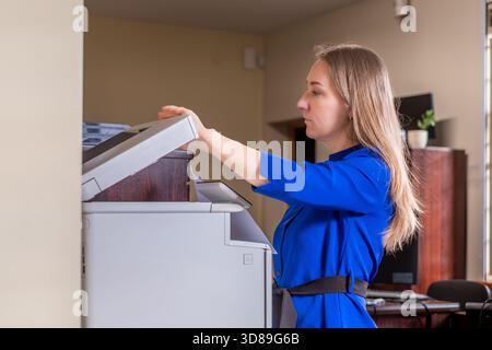 Female professional in blue shirt is using a printer in a contemporary office setting, highlighting business printing activities and workspace ambianc Stock Photo