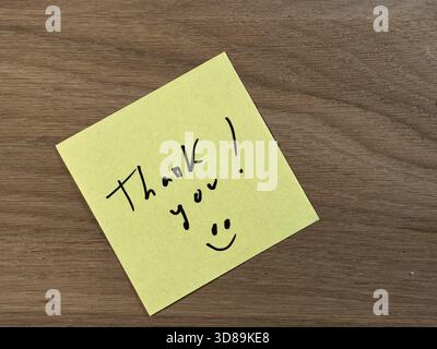 Handwritten Thank You on Yellow Sticky Note - Bright yellow sticky note with a handwritten 'Thank you!' message and smiley face on a warm, textured wo Stock Photo