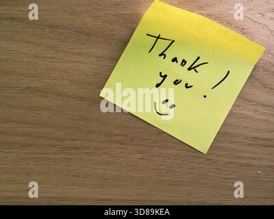 Handwritten Thank You on Yellow Sticky Note - Bright yellow sticky note with a handwritten 'Thank you!' message and smiley face on a warm, textured wo Stock Photo