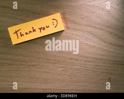 Thank You Note on Wooden Desk - A heartfelt yellow sticky note with "Thank you :)" handwritten on it, placed on a warm wooden desk. Conveys gratitude Stock Photo