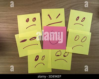 Happy Face Among Sad Sticky Notes - A single pink smiley face sticky note stands out from many yellow sad, angry, and worried faces on a wooden desk, Stock Photo