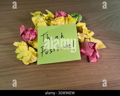 Think Positive Note on Crumpled Paper - An inspirational sticky note "Think Positive!" sits atop a pile of colorful crumpled papers on a wooden desk, Stock Photo