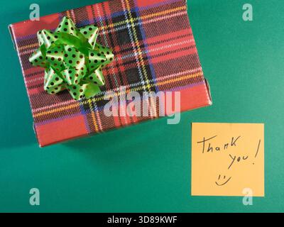 Plaid Gift with Thank You Note - Overhead view of a red plaid gift box with a green polka-dot bow, accompanied by a yellow sticky note expressing grat Stock Photo