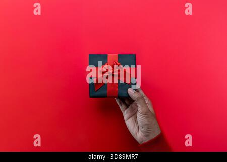 Top view image of hand holds a black gift box with a red ribbon and bow against a vibrant red background. Gift-giving, holidays, sales events Stock Photo