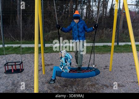 Two brothers playing on nest swing at playground with older boy standing and pushing younger sibling on autumn evening. Concept of siblings outdoor fu Stock Photo