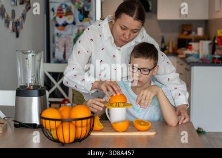 Mother guiding son hands while squeezing orange juice together with manual juicer in home kitchen. Concept of parent teaching cooking skills, family b Stock Photo