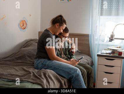 Mother and son sitting together on bed looking at smartphone in cozy kids room with wall stickers. Concept of family screen time, digital parenting an Stock Photo
